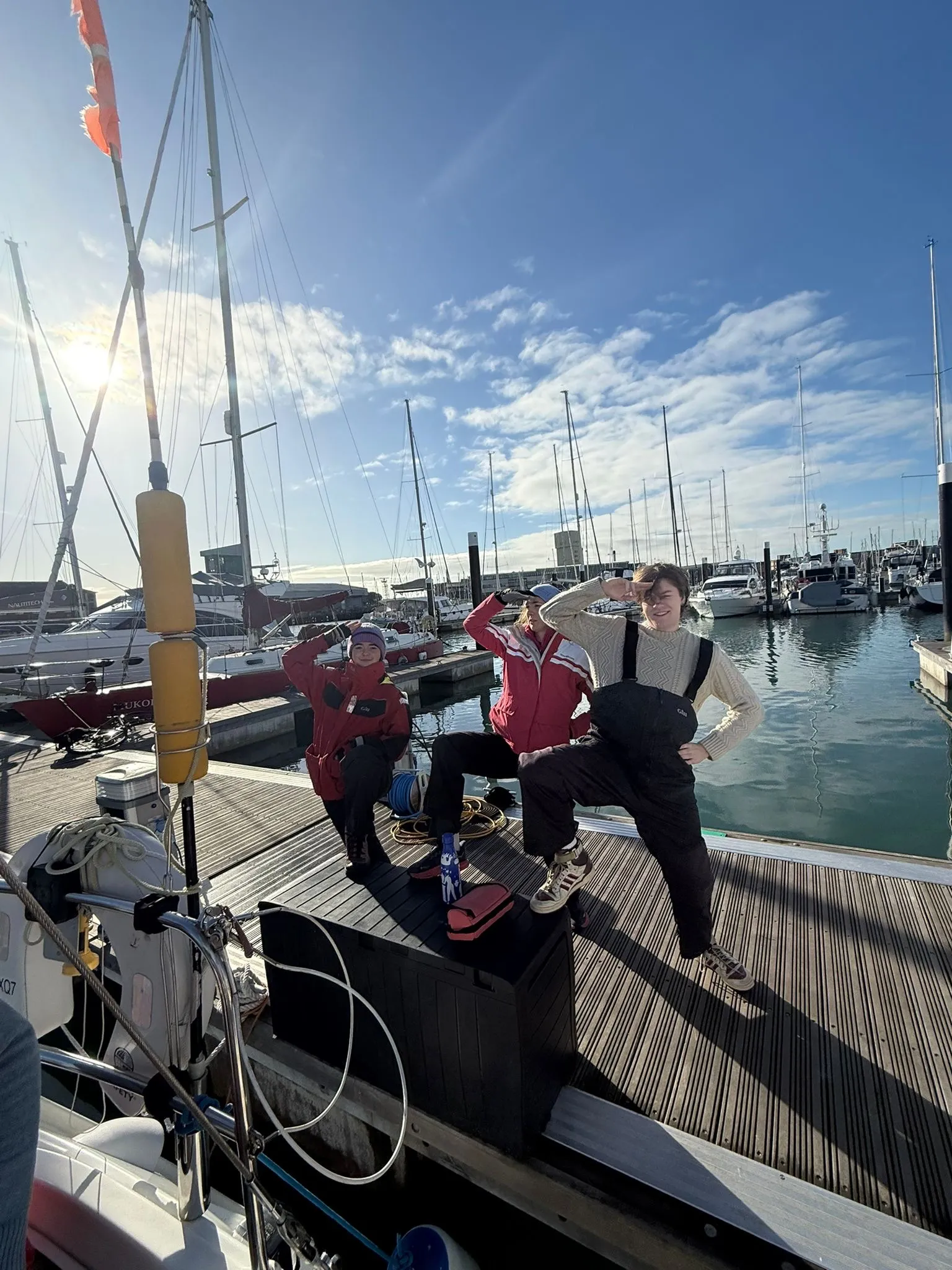 Three people posing playfully on a dock in front of boats and yachts under a sunny blue sky.