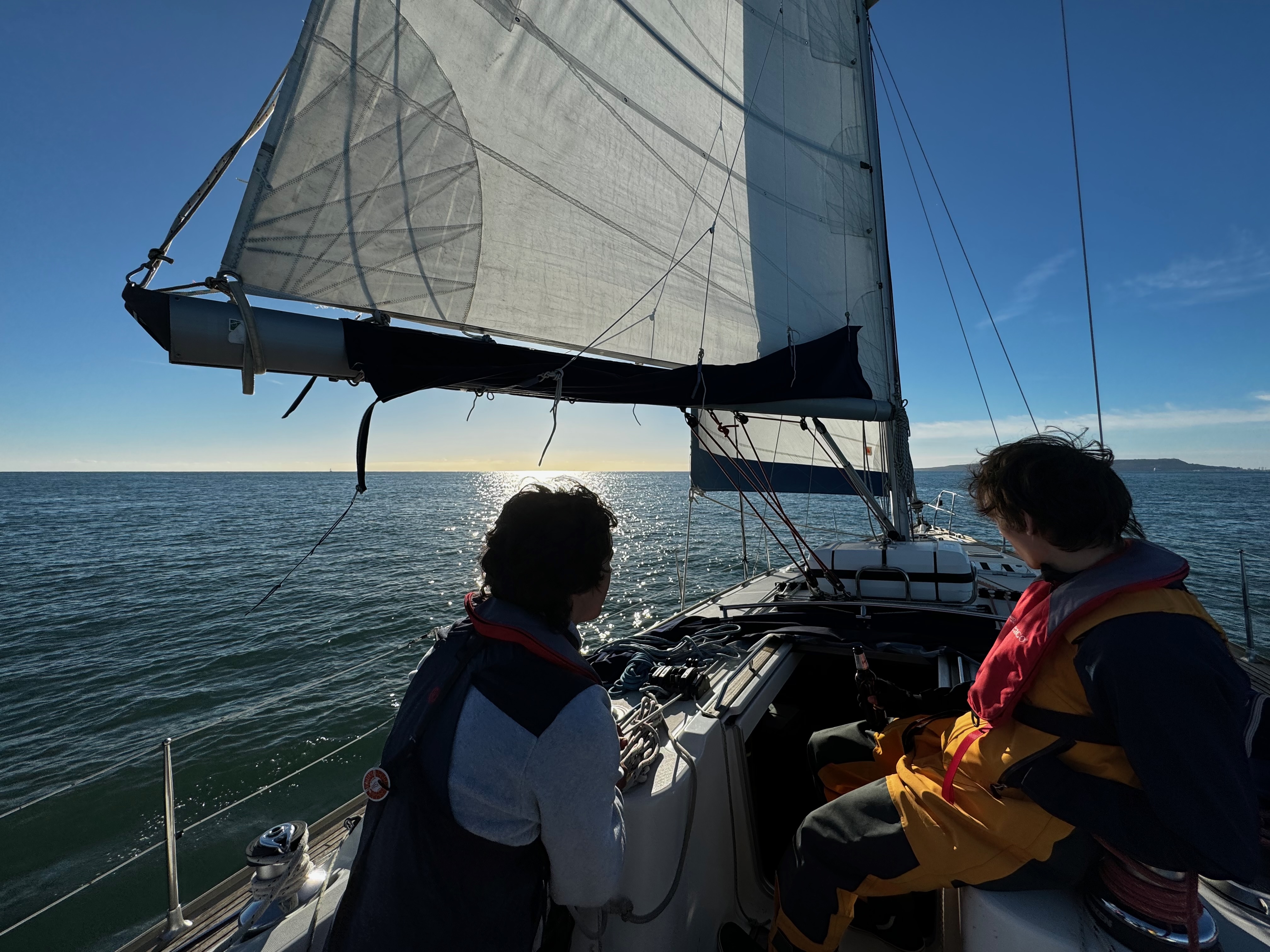 Two people wearing life jackets sailing on a sailboat with the sun reflecting on the calm sea.