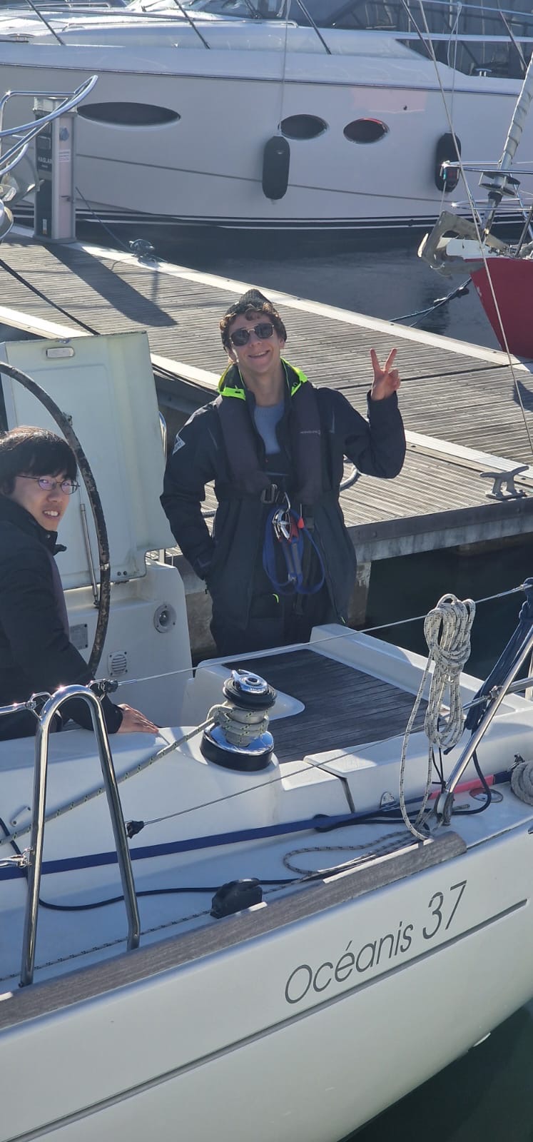 Two young men on a sailboat named Océanis 37 docked at a marina, one smiling and giving a peace sign.