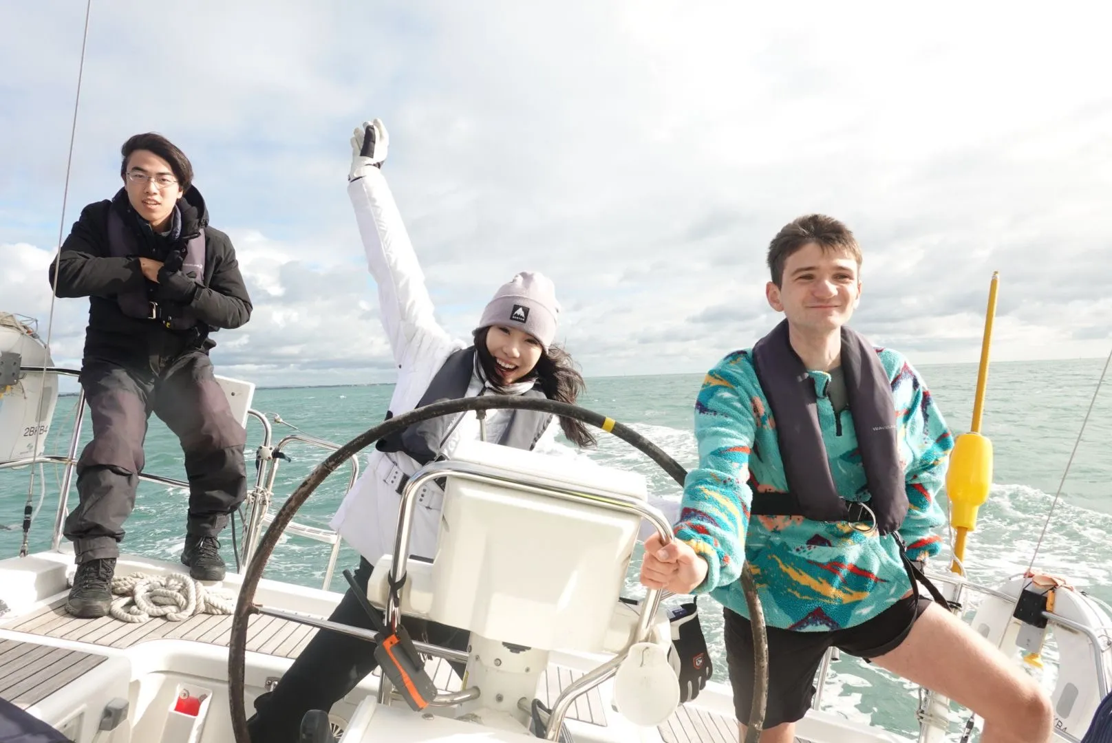 Three people enjoying sailing on a boat, with one person steering the wheel, another raising an arm cheerfully, and a third standing nearby in life jackets.