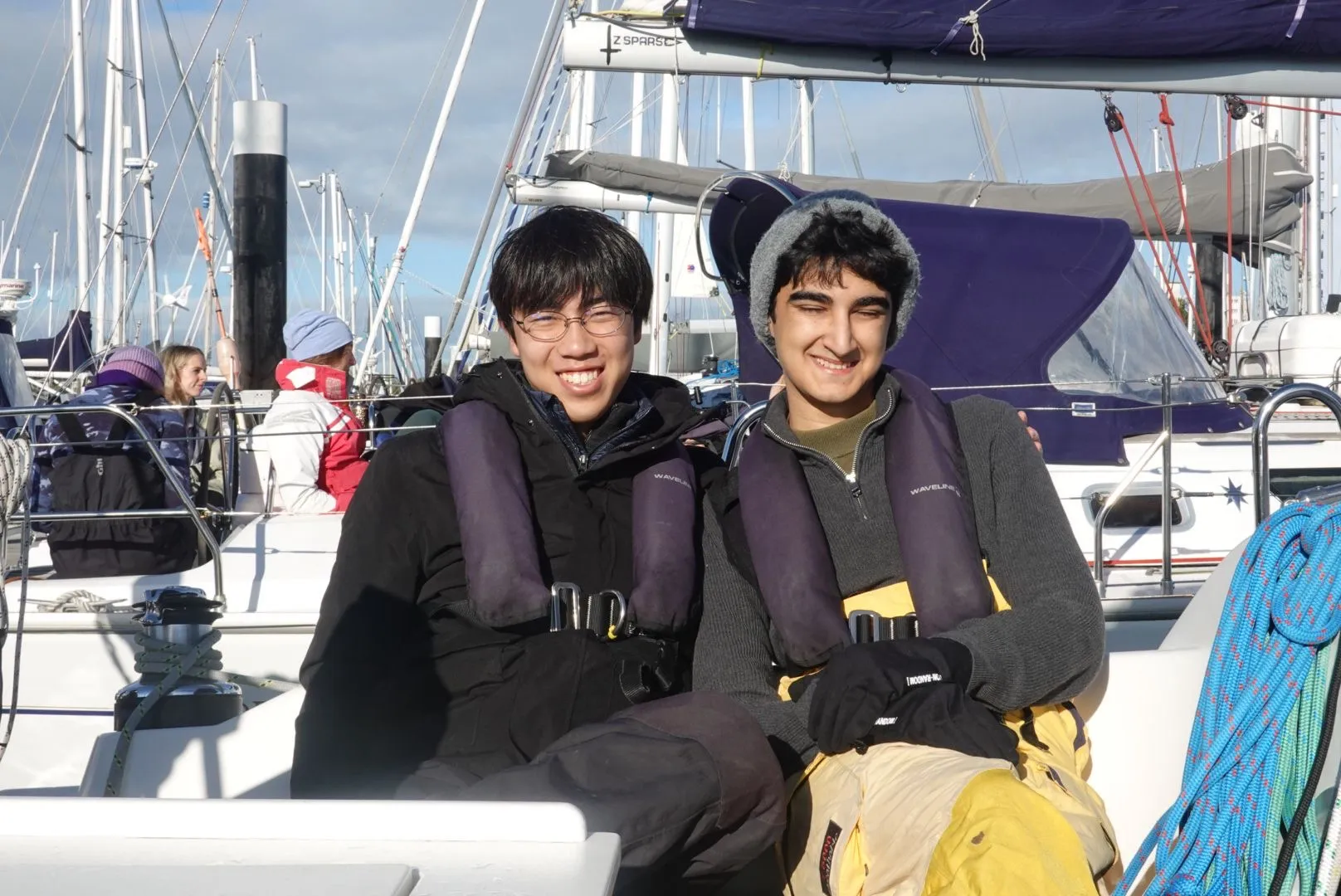 Two young men smiling and wearing life jackets while sitting on a boat with other boats and people in the background.
