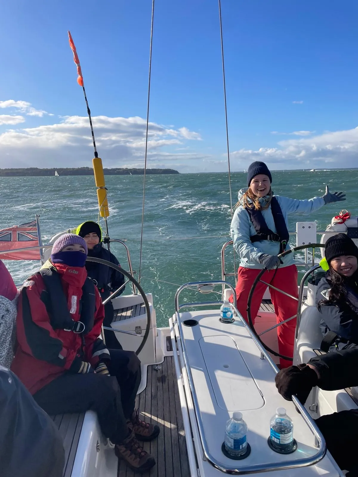 Four people dressed in warm clothes and life jackets on a sailboat steering with two wheels under a blue sky and choppy sea.