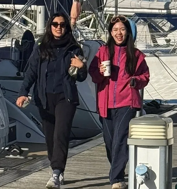 Two women walking on a wooden dock near boats, one holding a coffee cup and the other wearing sunglasses.