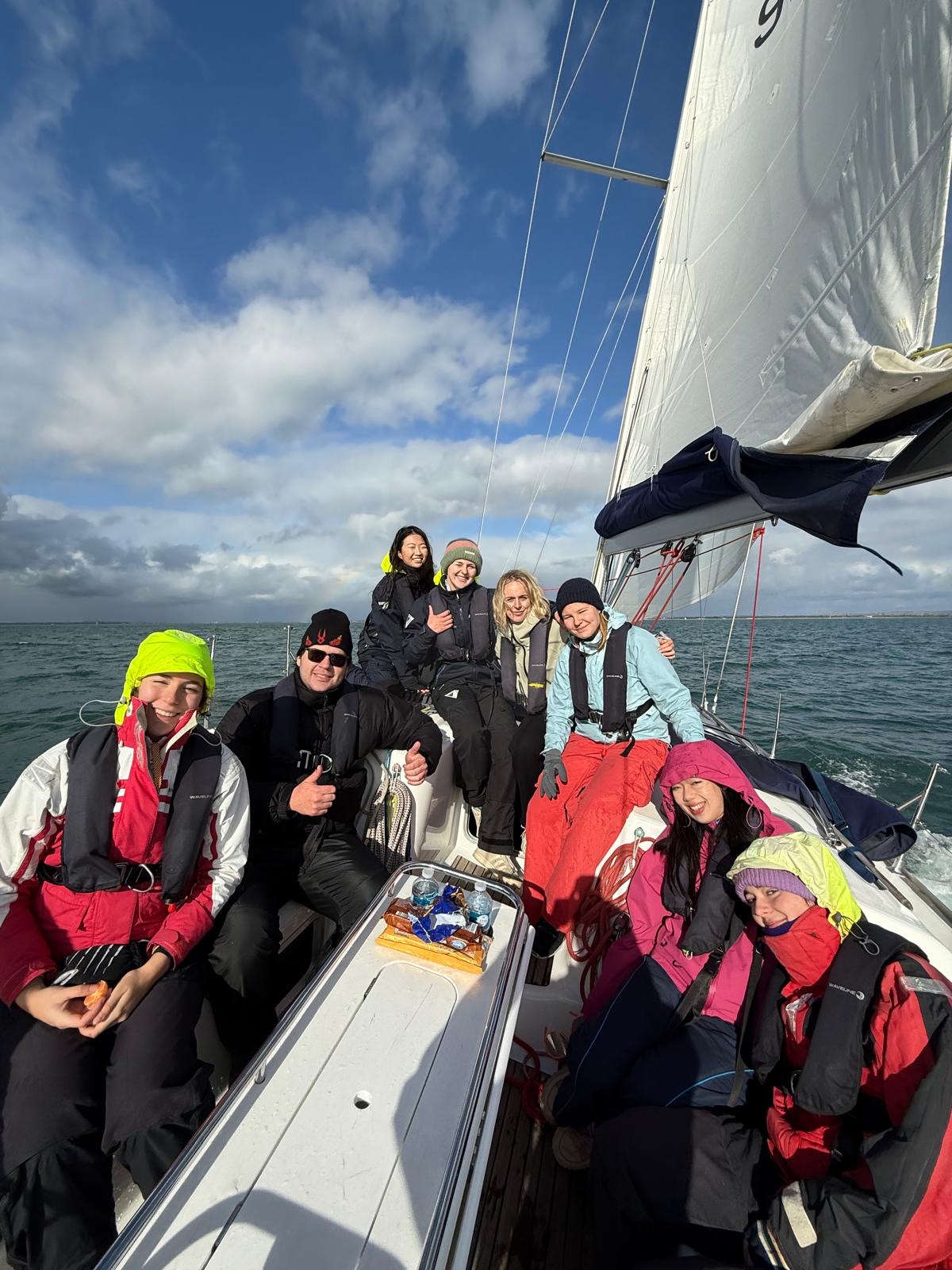 Seven people in warm clothing and life jackets smiling and sitting together on a sailboat under a partly cloudy blue sky.