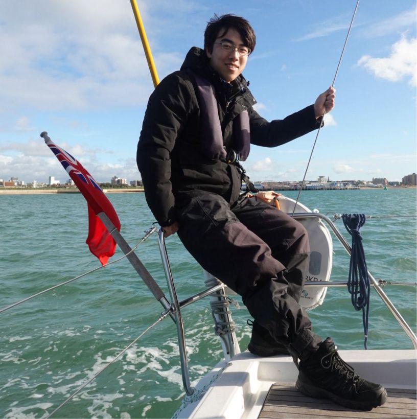 Smiling man in black jacket and life vest sitting on the edge of a sailboat with a red flag, blue sky, and coastal city in the background.