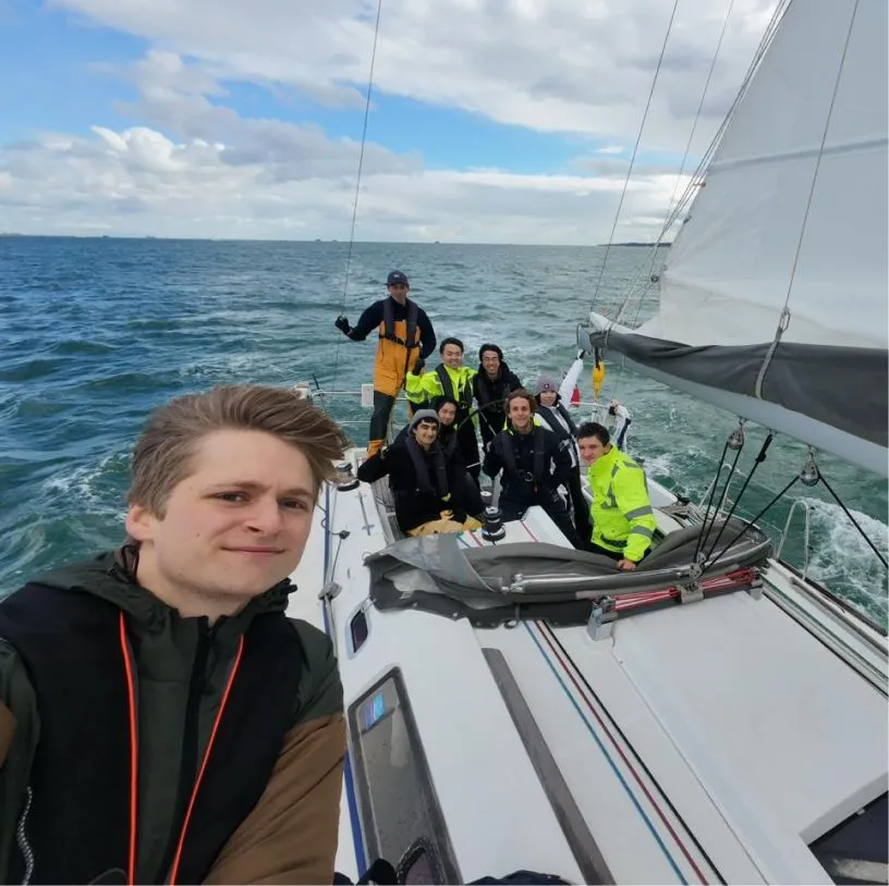 Group of eight people wearing life jackets and sailing gear on a sailboat in the ocean under a partly cloudy sky.