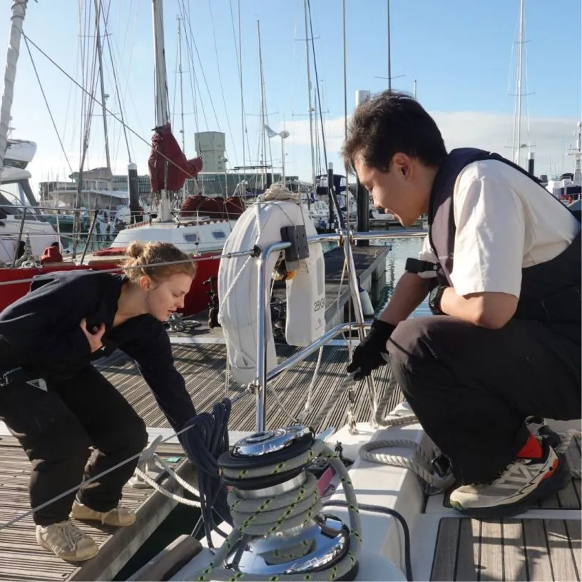 Two people working with ropes on a docked sailboat at a marina on a sunny day.