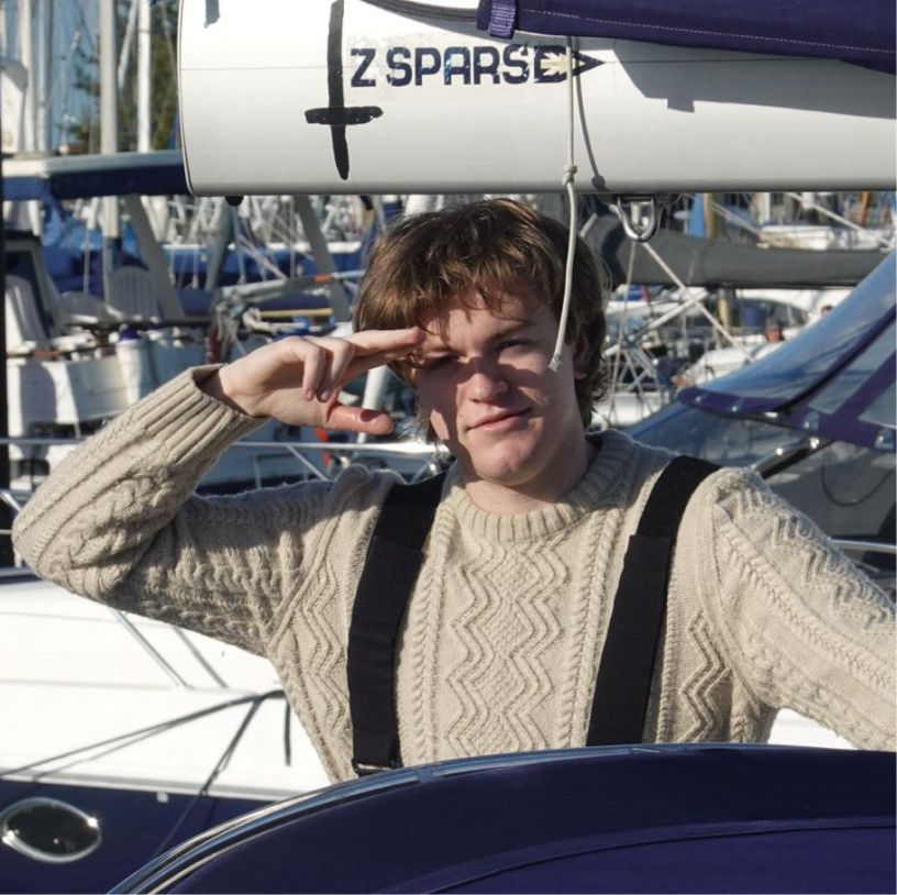 Young man in a beige cable-knit sweater saluting while standing on a boat at a marina.