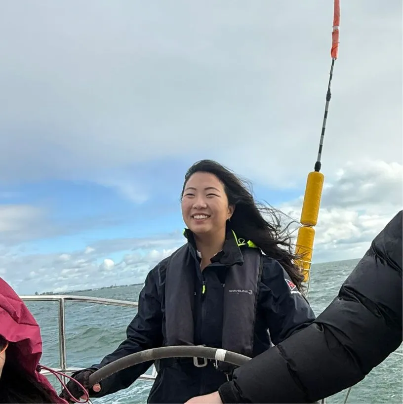 Smiling woman steering a boat on a windy day with ocean and cloudy sky in the background.