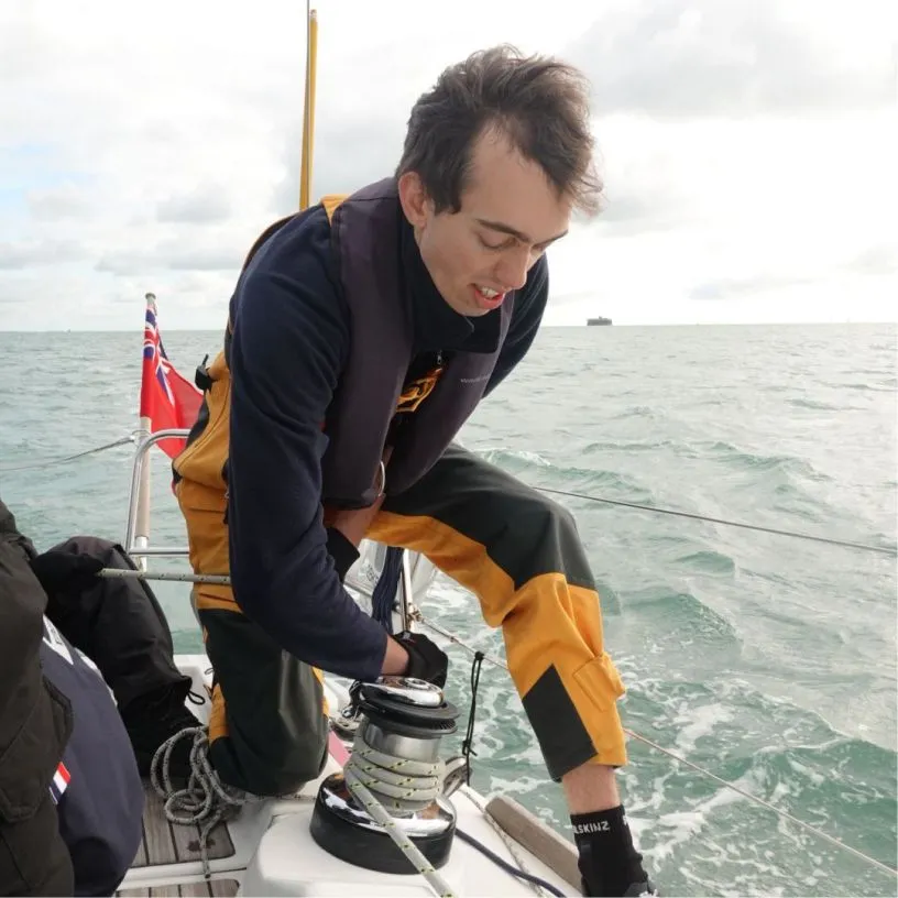 Man wearing yellow and black waterproof pants adjusting a winch on a sailboat with ocean in the background.