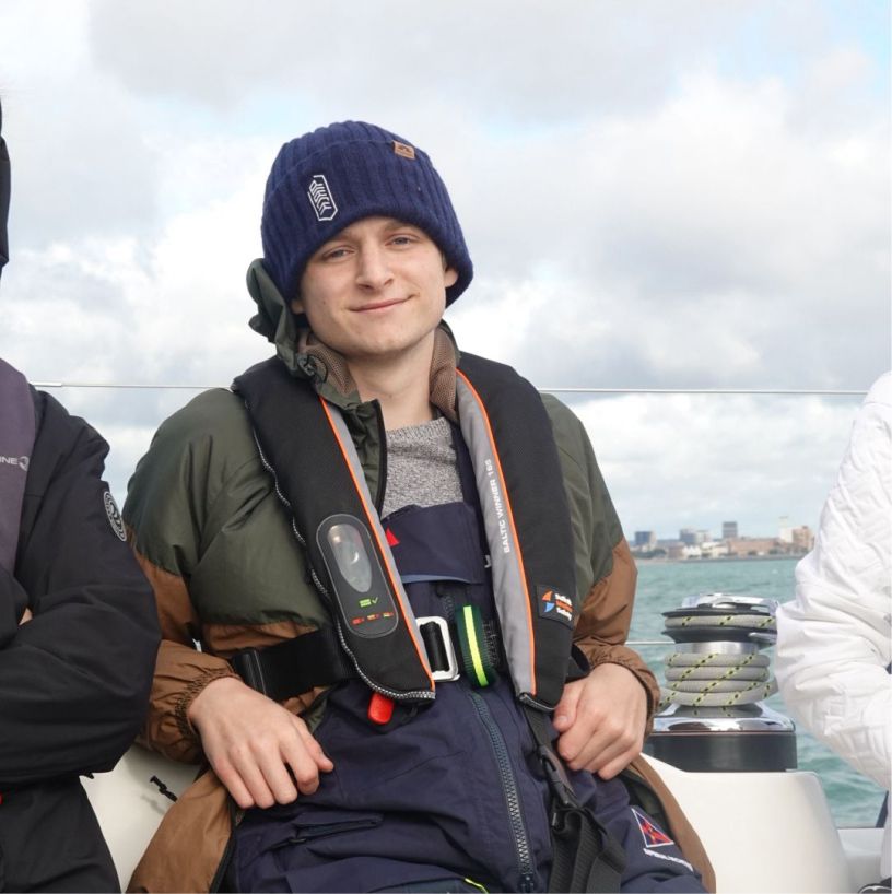 Young man wearing a blue knit hat and life jacket sitting on a boat with water and cityscape in the background.