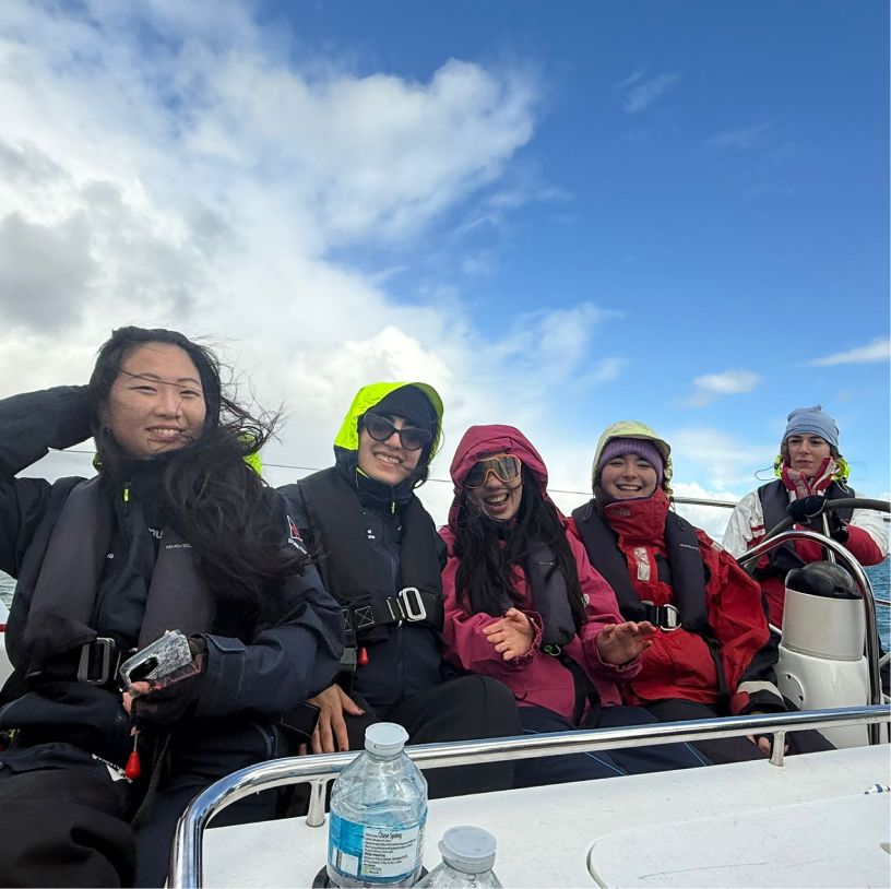 Five people wearing life jackets and warm jackets smiling and sitting on a boat under a partly cloudy blue sky.