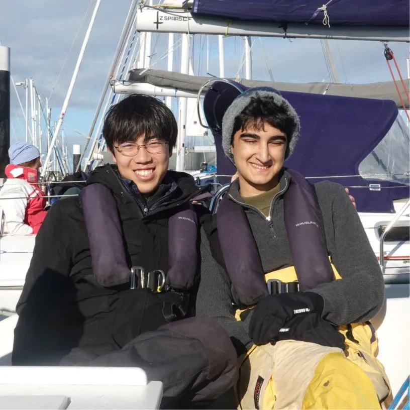 Two young men smiling and wearing life jackets sitting on a sailboat with masts and rigging in the background.