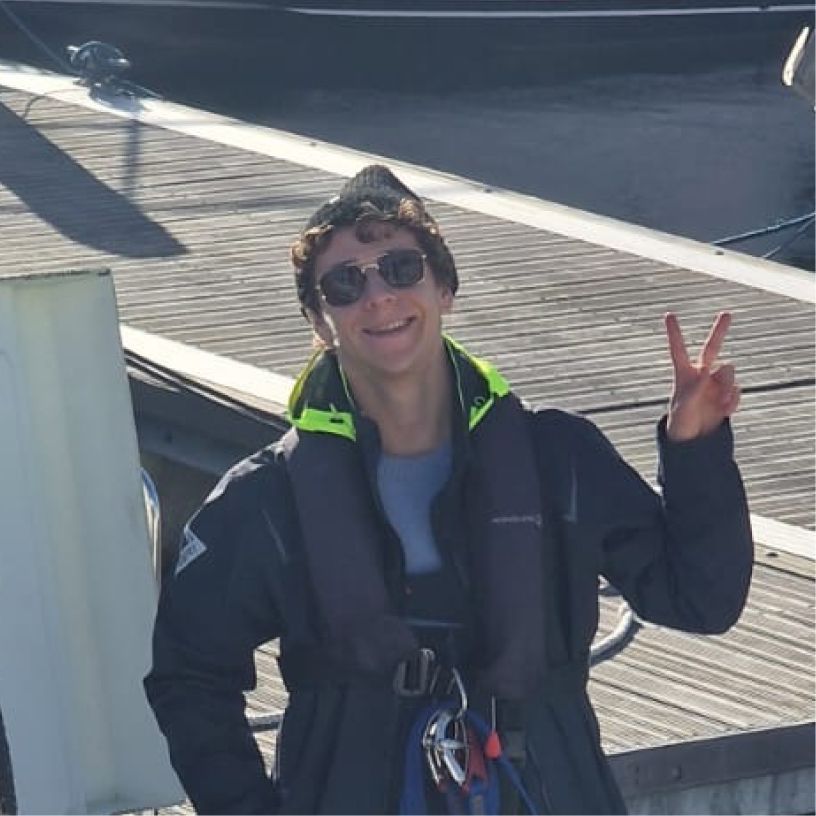 Young man wearing sunglasses and a life jacket standing on a dock, flashing a peace sign and smiling.