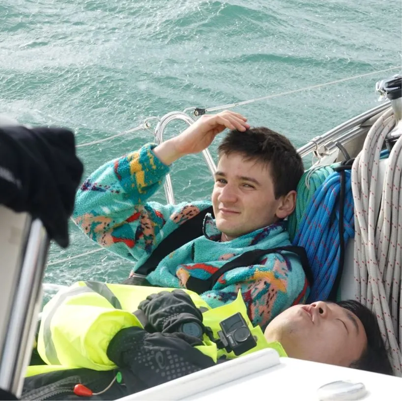 Two young men relaxing on a sailboat near coiled ropes with water in the background, one smiling and adjusting his hair.