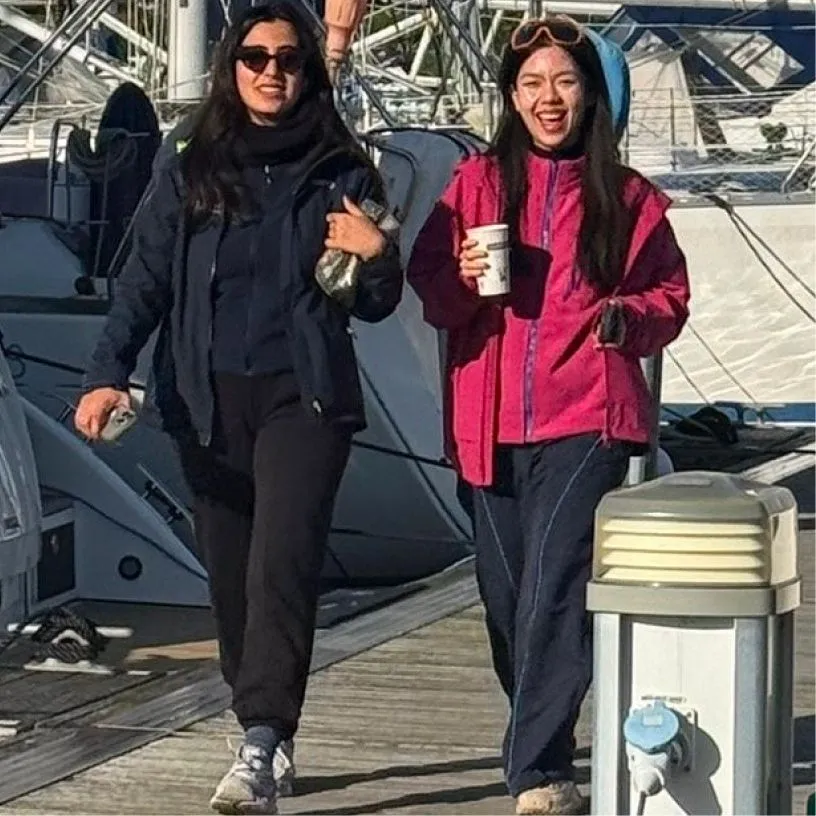 Two women walking on a dock beside boats, one holding a cup and smiling, the other wearing sunglasses.
