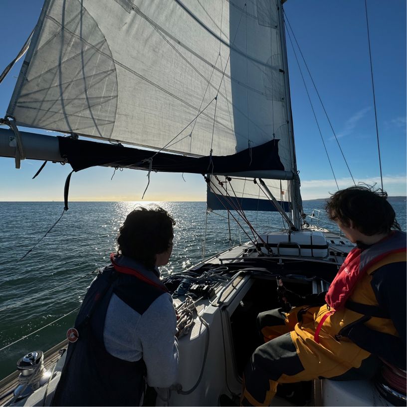 Two people wearing life jackets on a sailboat, facing the ocean under a large sail with the sun reflecting on the water.