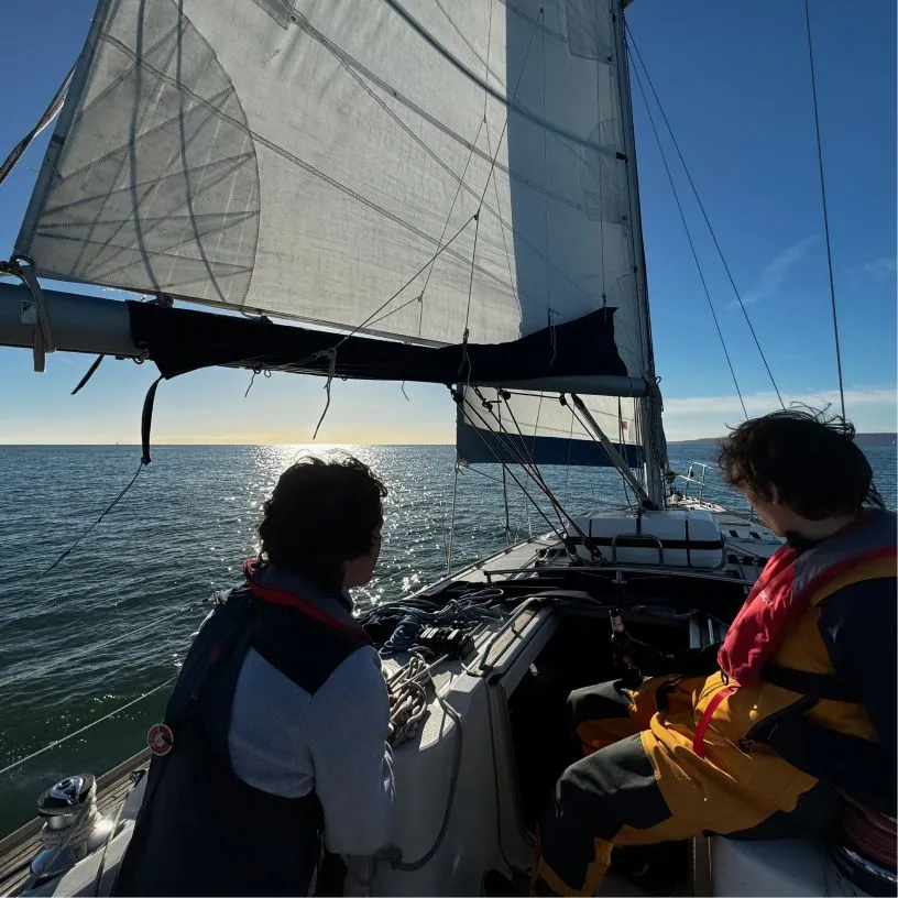 Two people wearing life jackets on a sailboat, facing the ocean under a large sail with the sun reflecting on the water.