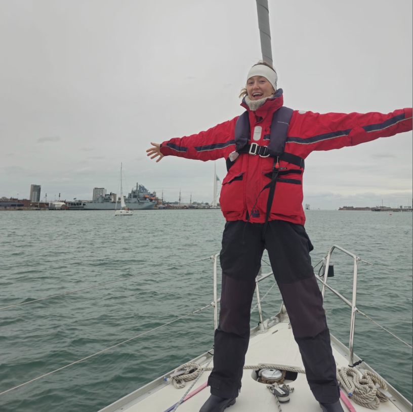 Smiling person in red jacket and headband standing on a sailboat with arms outstretched over the water near a harbor city.