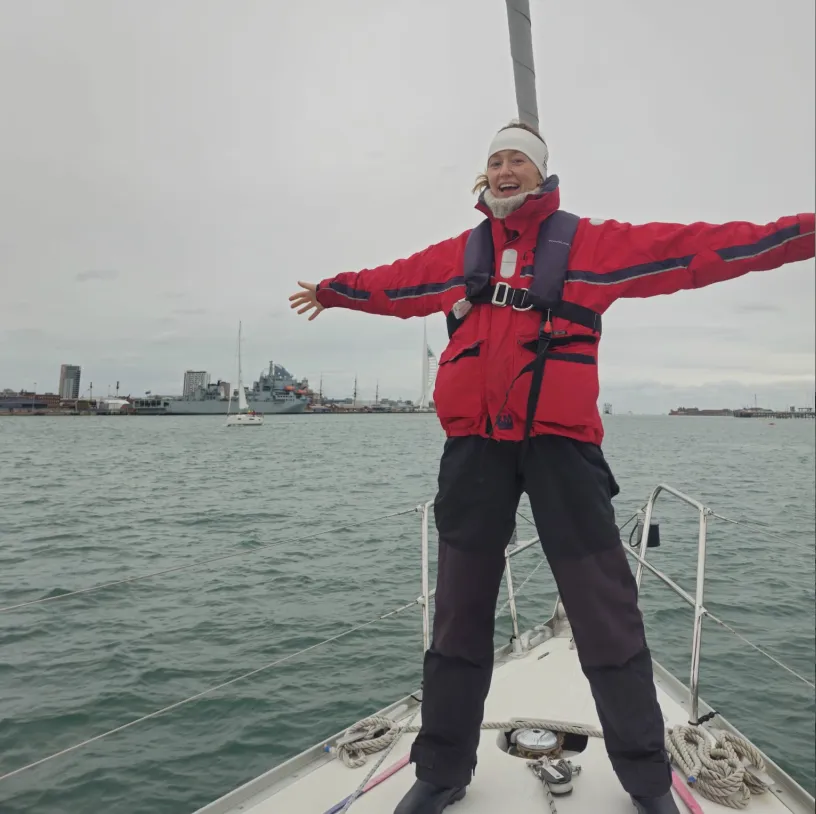 Smiling person in red jacket and headband standing on a sailboat with arms outstretched over the water near a harbor city.
