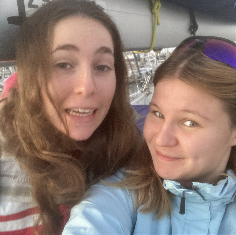 Two young women smiling, one wearing a light blue jacket and sunglasses on her head, with boats visible in the background.