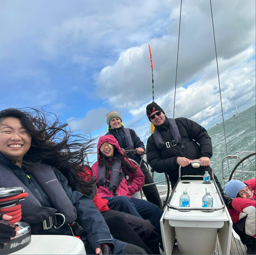 Five people wearing jackets and life vests smiling and enjoying a windy day on a sailboat with cloudy skies and choppy water.
