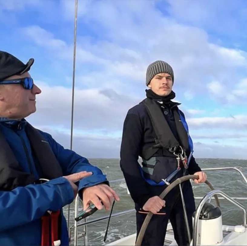 Two men on a sailboat, one steering the wheel wearing a knit hat and life vest, with water and cloudy sky in the background.