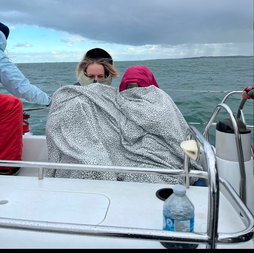 Two people wrapped in matching leopard-print blankets sitting close together on a boat with choppy ocean and cloudy sky in the background.