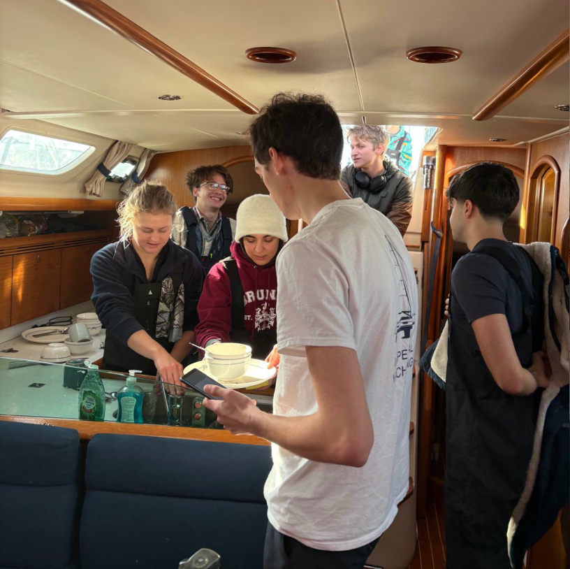 Group of six young people socializing inside a boat cabin with wooden interior and kitchen area.