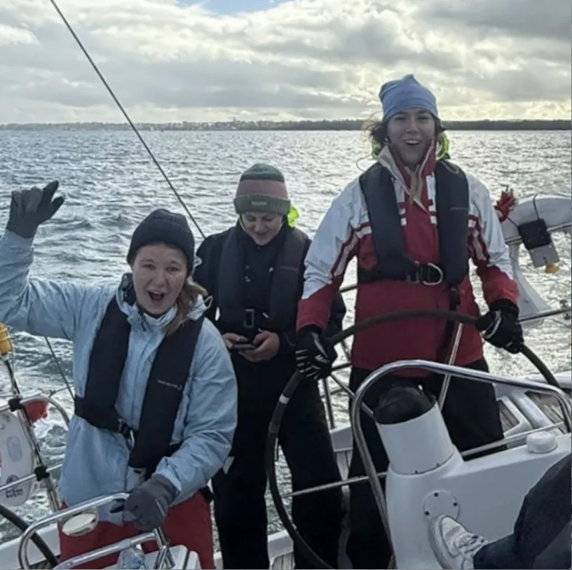 Three people wearing life jackets and warm clothing aboard a sailboat on the water, one waving and another steering the wheel.