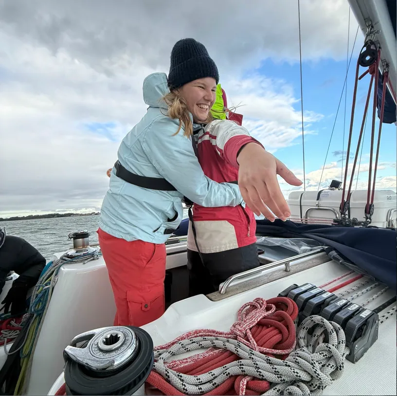 Two people on a sailboat, one wearing a light blue jacket and black beanie joyfully hugging another in a red and white jacket, with ropes and sailing equipment visible.