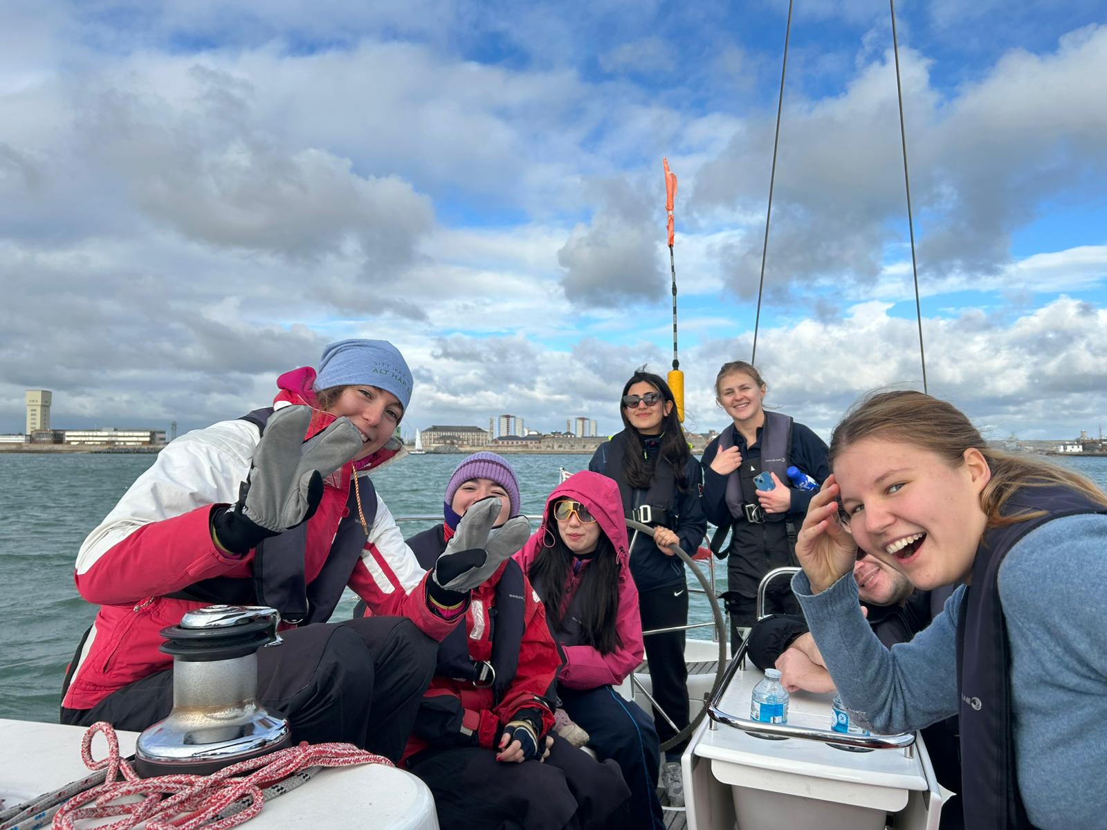 Group of six people on a sailboat smiling and enjoying a partly cloudy day on the water.