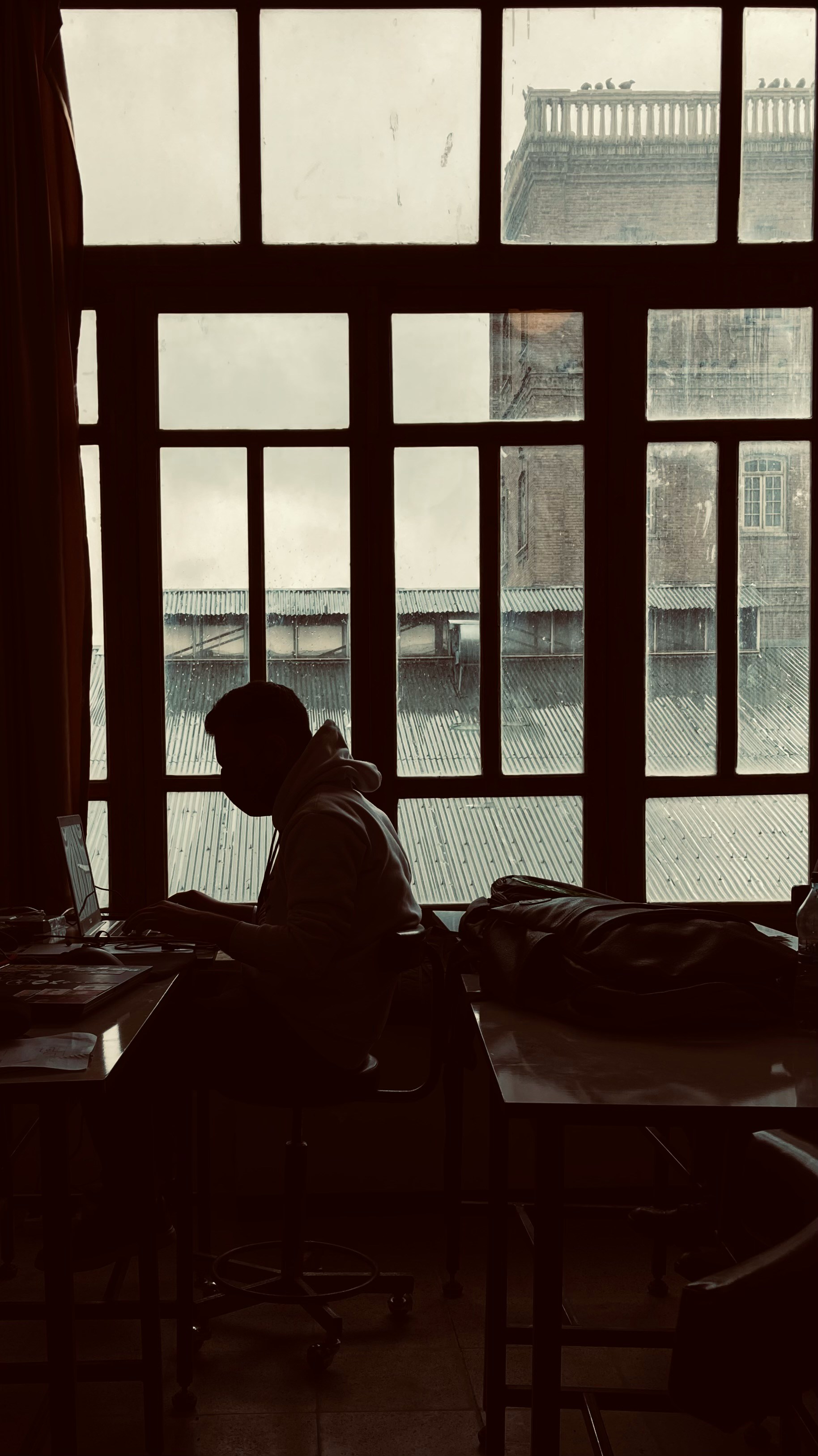 Silhouette of a person sitting and working on a laptop in front of a large window with a view of a rooftop and building outside.