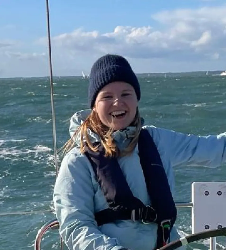 Smiling person wearing a blue jacket and black beanie steering a sailboat on choppy water under a partly cloudy sky.