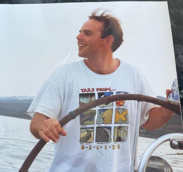 Man steering a boat with a large wooden wheel, smiling and wearing a white T-shirt with colorful animal footprints labeled 'Tails from Belize.'