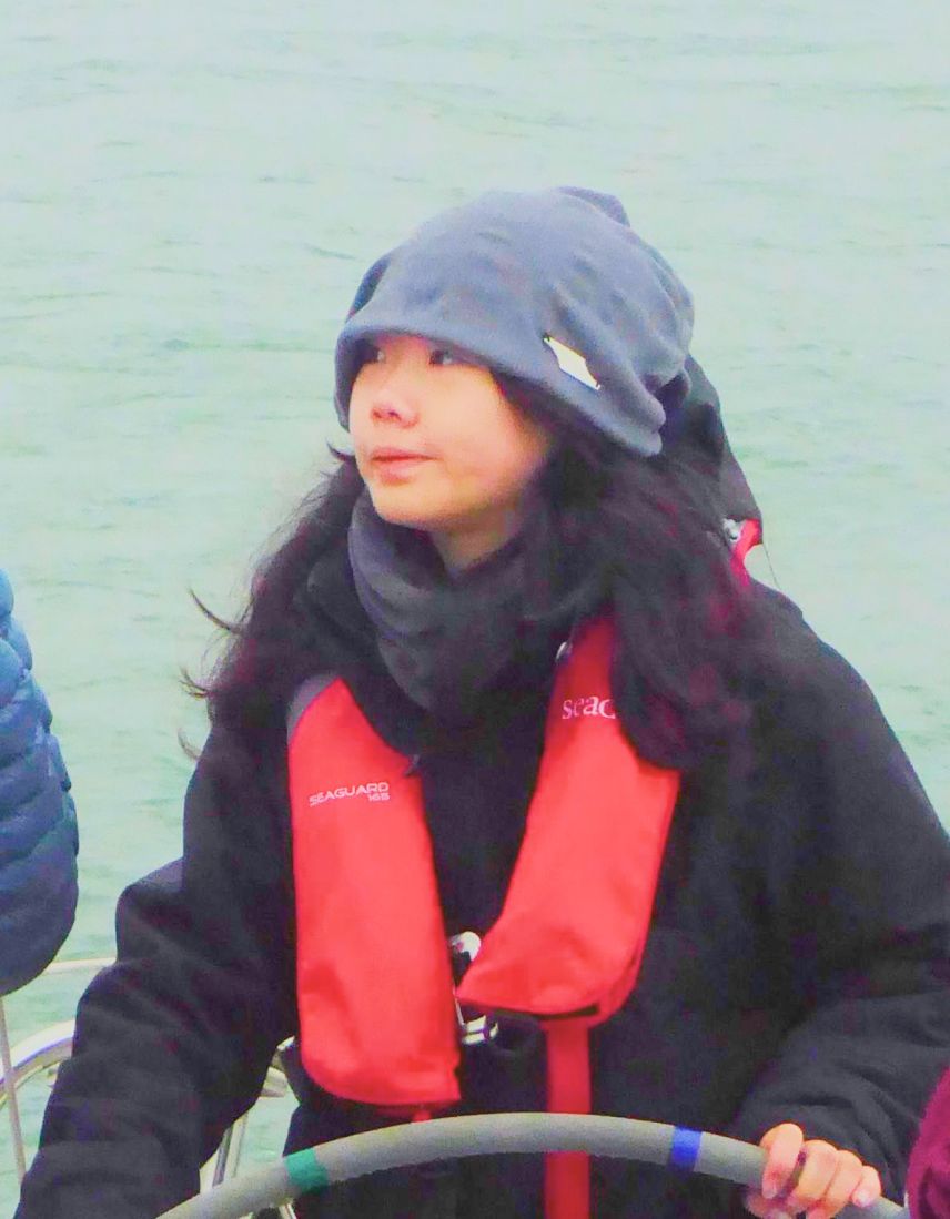 Person wearing a gray beanie and red life jacket steering a boat with water in the background.