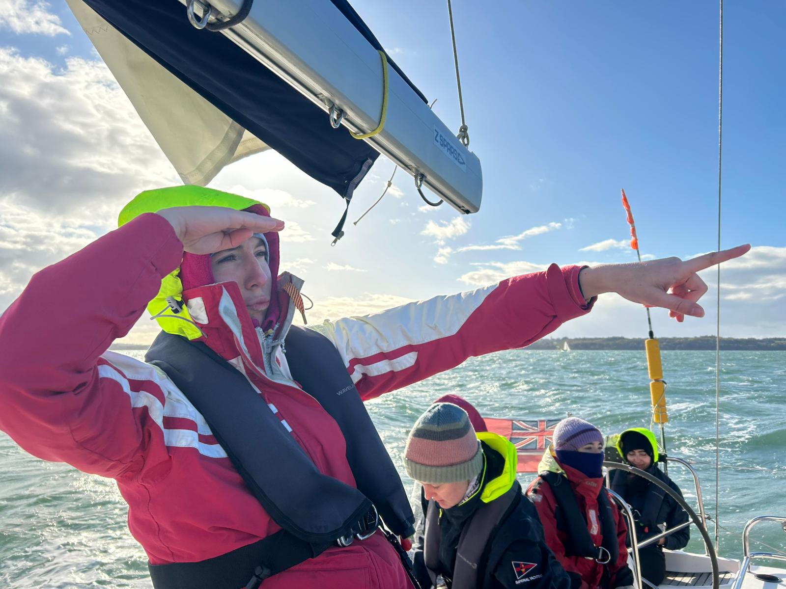Person in red jacket and life vest on boat pointing forward while shading eyes, with three other people in warm clothing sitting behind.