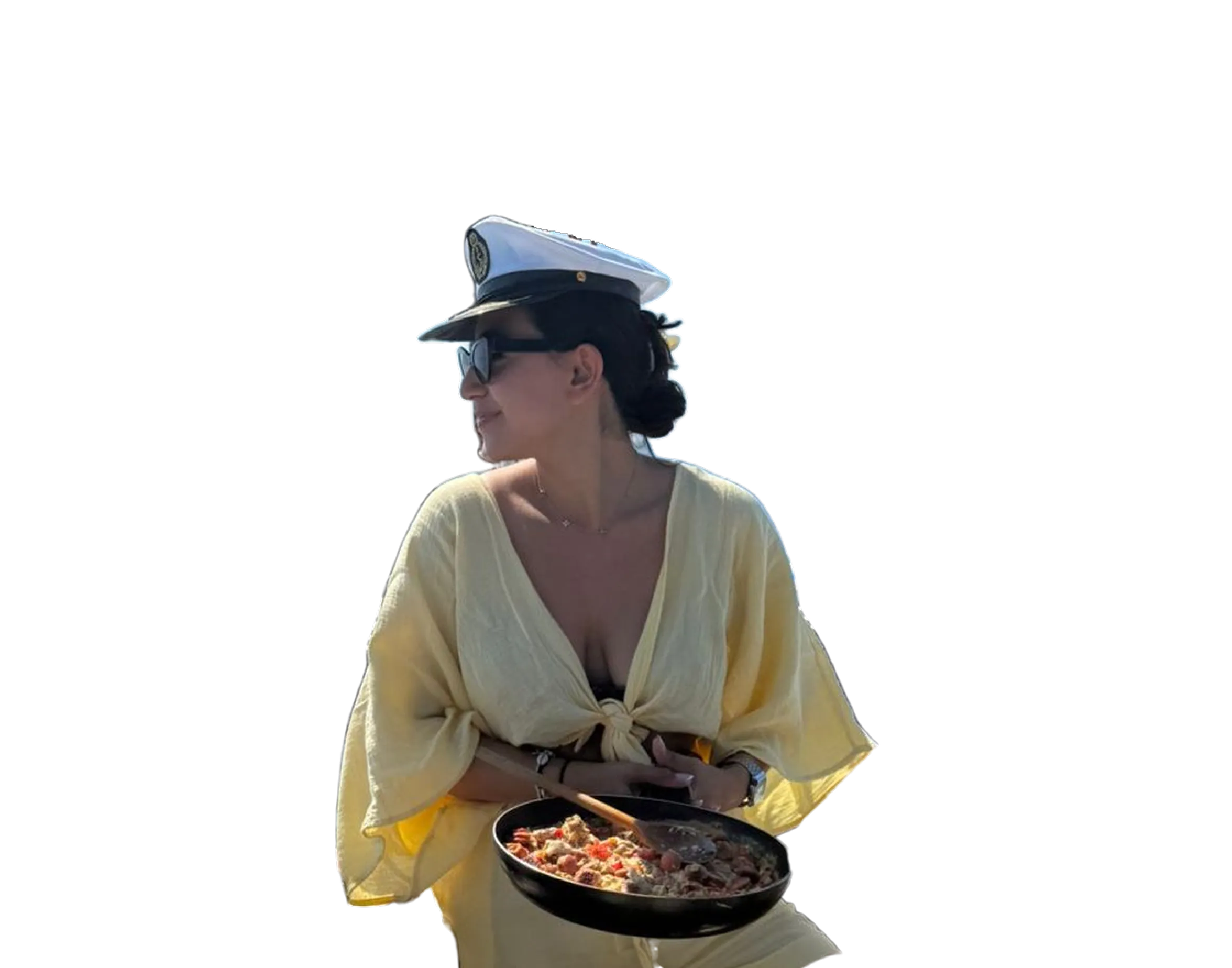 Woman in a light yellow dress and white captain hat holding a pan of cooked food with a wooden spoon.