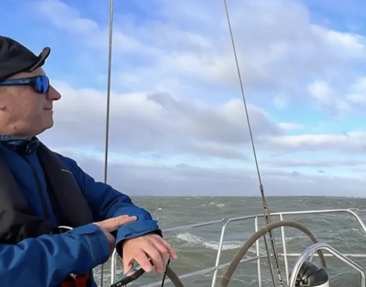 Man wearing sunglasses and a hat steering a sailboat on a cloudy day with windy waters.