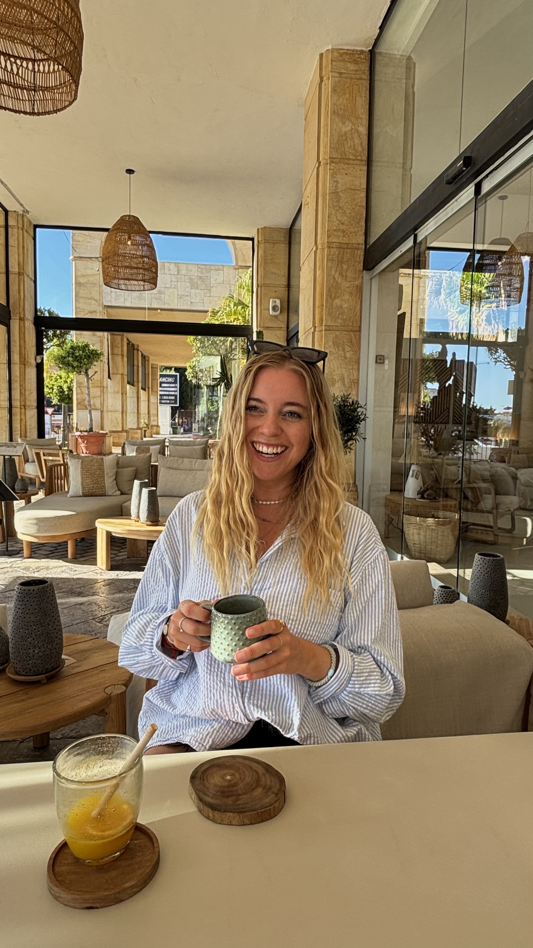 Smiling woman with long blonde hair holding a green textured mug, seated at a table with a glass of orange juice and wooden coasters in a sunlit outdoor patio.