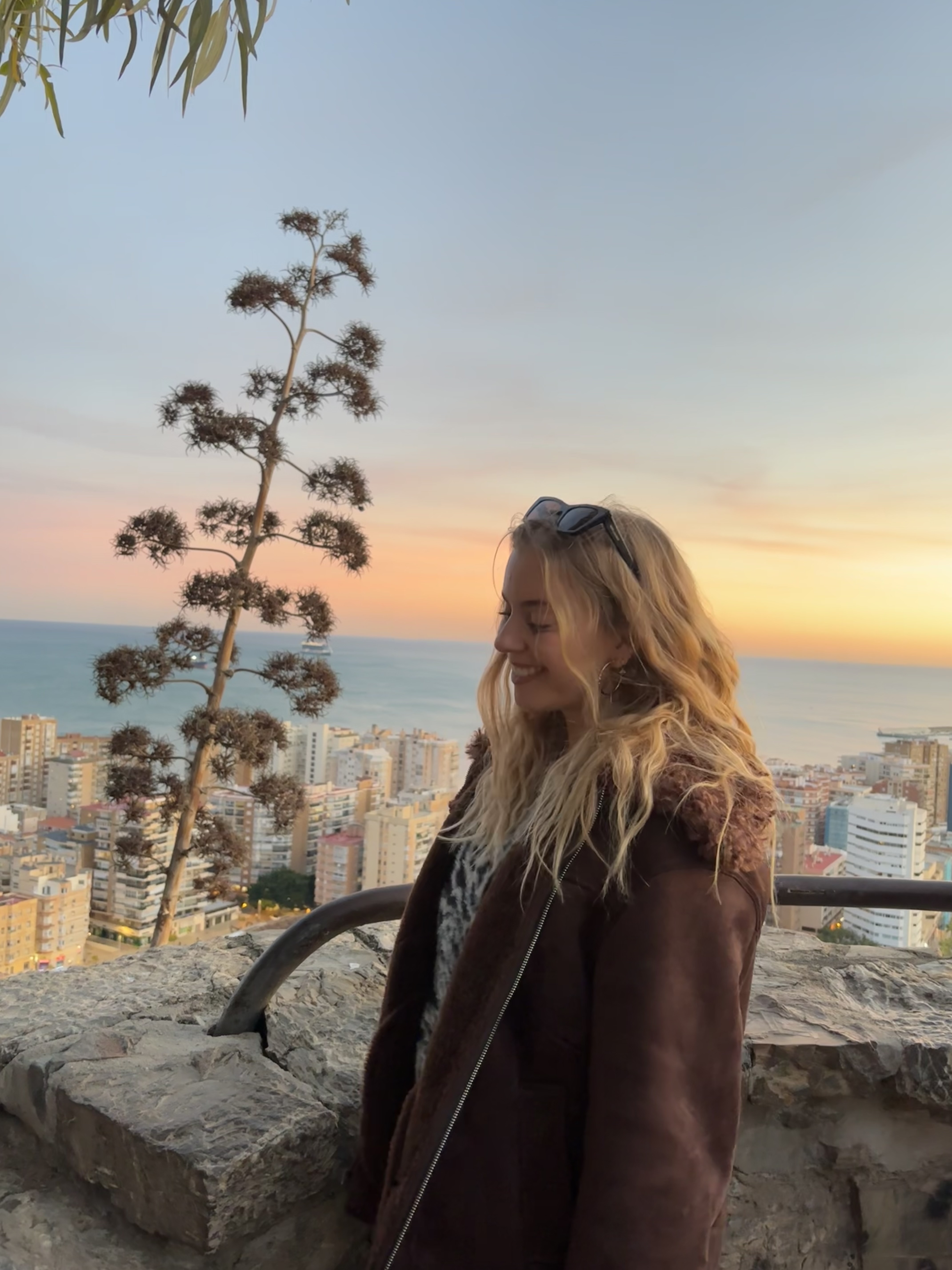 Smiling woman with long blonde hair wearing a brown coat, standing on a stone terrace overlooking a coastal city at sunset.