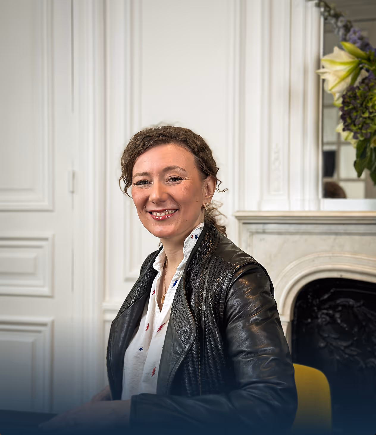 Portrait d'une femme souriante portant une veste en cuir noir et une chemise blanche avec des motifs colorés, assise devant une cheminée ornée.