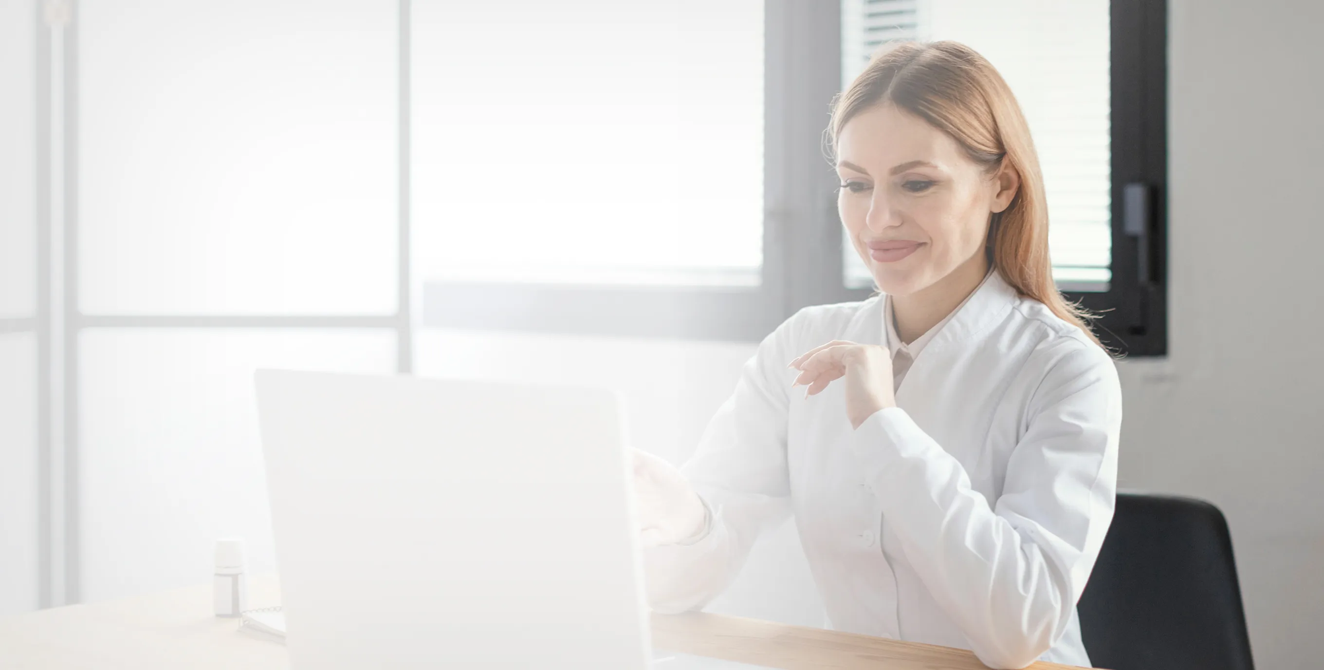 Smiling female doctor in white coat sitting at a desk using a laptop.