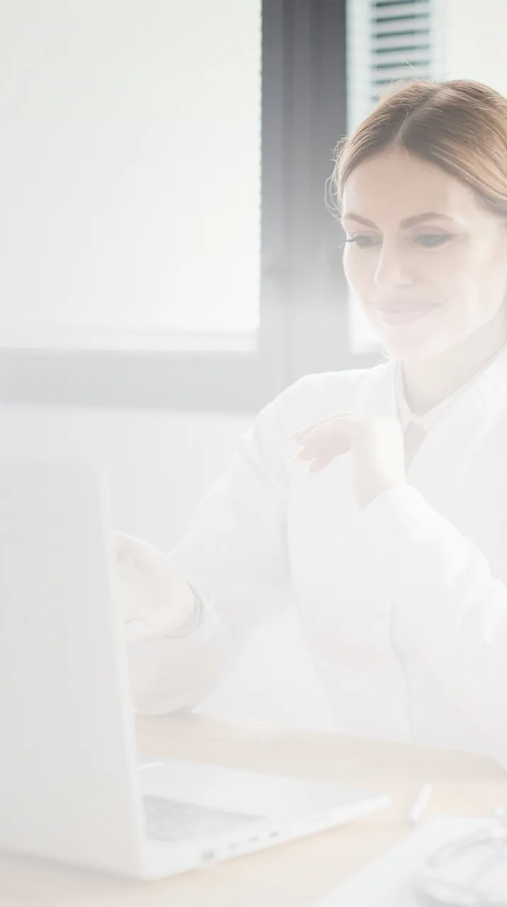 Smiling woman in white shirt working on a laptop at a wooden desk near a window.