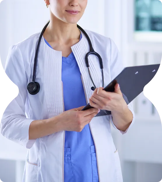 Female doctor in white coat and blue scrubs holding a clipboard with a stethoscope around her neck.