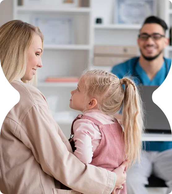 Mother holding her young daughter while a male doctor in scrubs smiles in the background during a medical consultation.