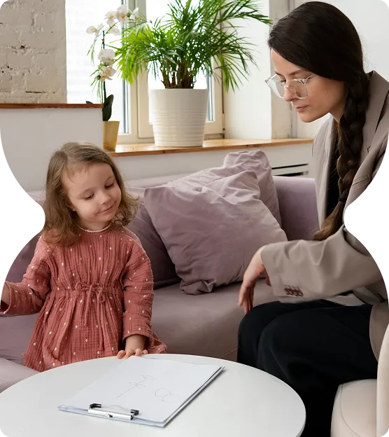 Young girl in a pink dress sitting on a couch next to a woman with glasses, both looking at a clipboard with drawings on a white table.
