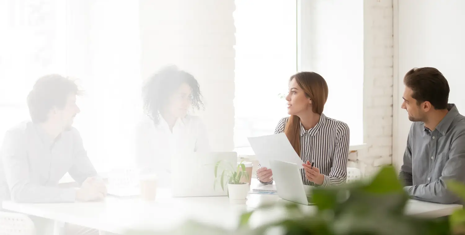 Four colleagues having a discussion in a bright office around a white table with laptops and papers.