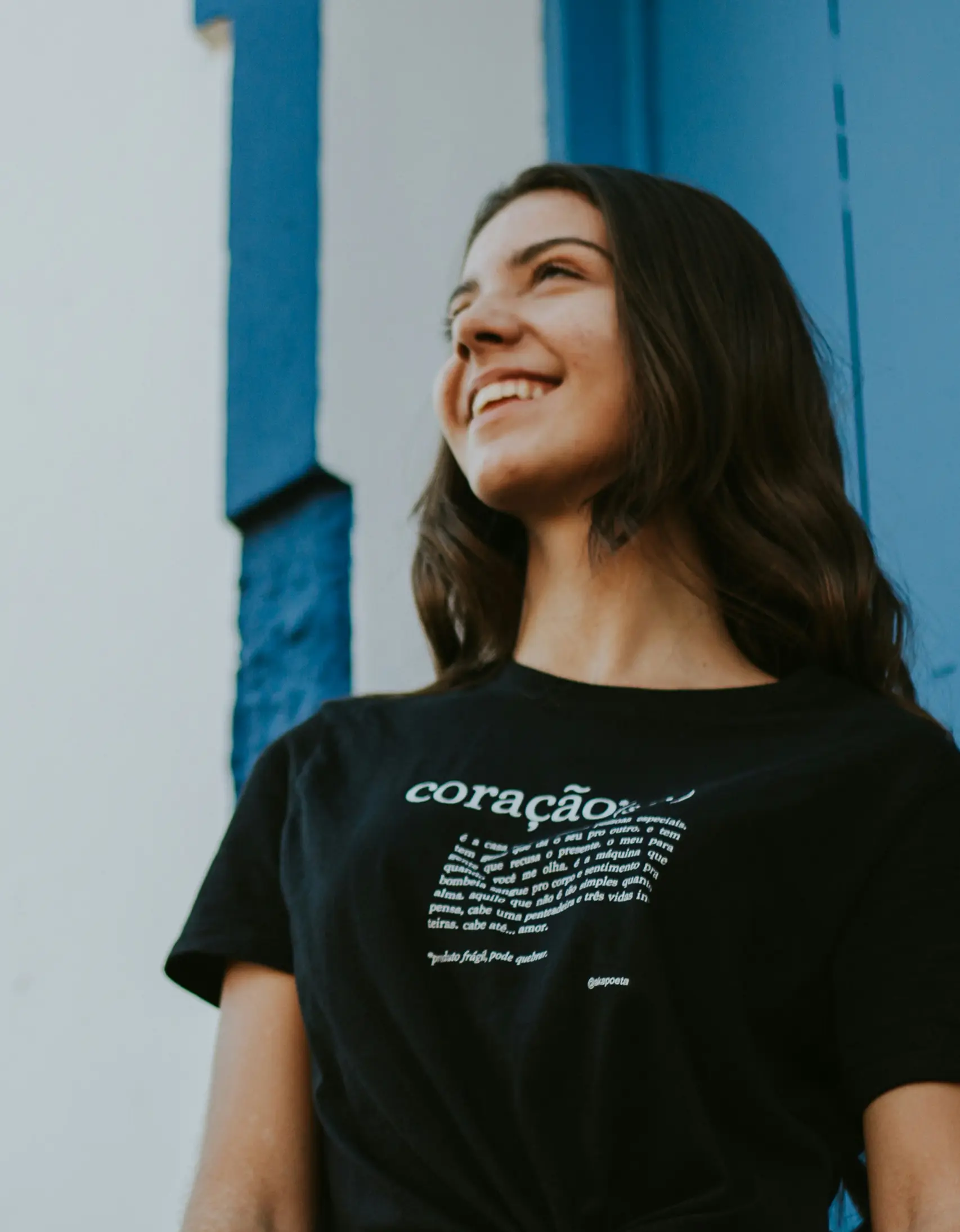 Smiling young woman with long dark hair wearing a black t-shirt with white text, standing against a blue and white wall.