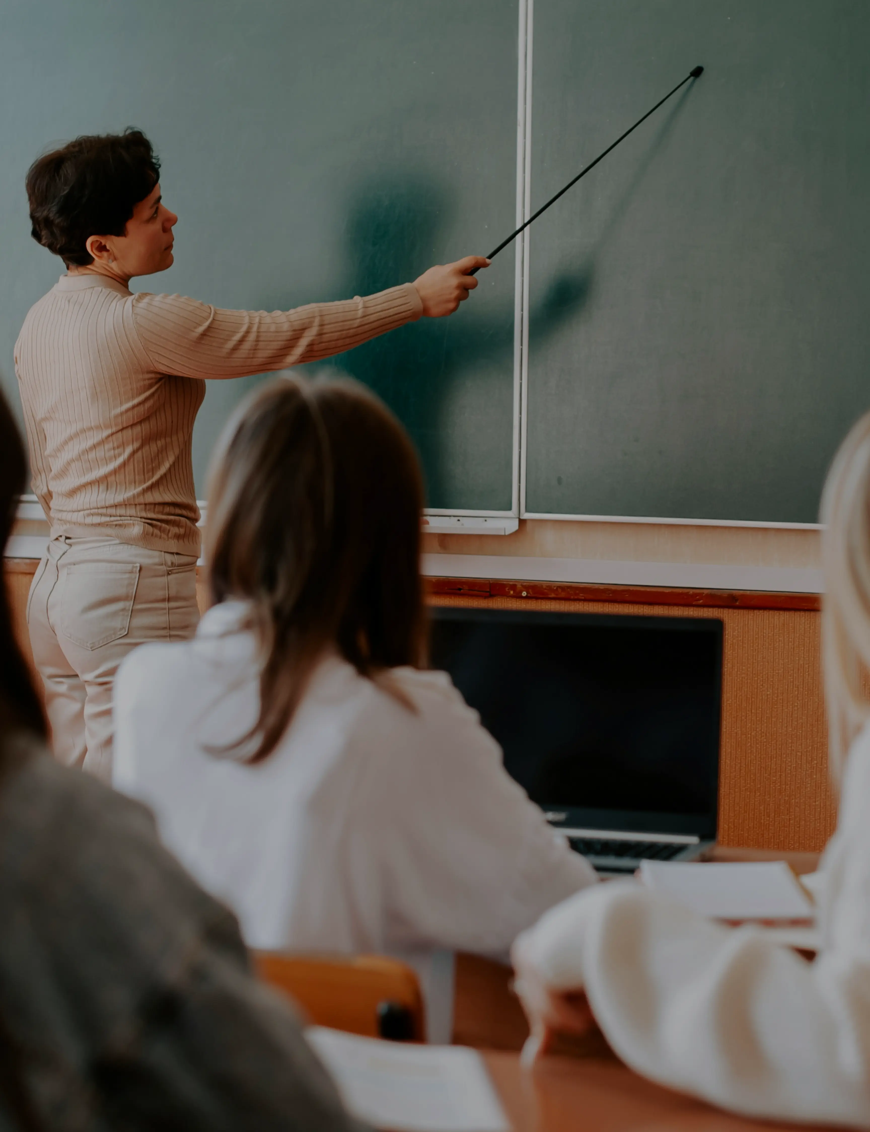 Teacher pointing at a blank chalkboard with a stick while students sit at desks with a laptop in a classroom.